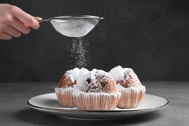 Photo of Woman with sieve sprinkling powdered sugar onto muffins at grey textured table, closeup
