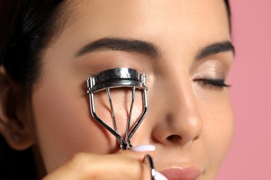 Young woman using eyelash curler on light pink background, closeup