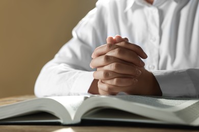 Woman holding hands clasped while praying over Bible at wooden table, closeup