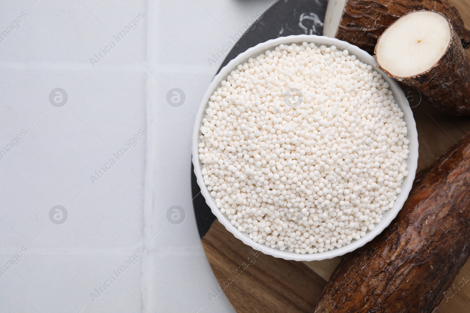 Photo of Tapioca pearls in bowl and cassava roots on white tiled table, flat lay. Space for text