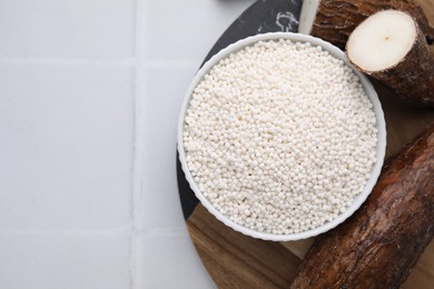 Tapioca pearls in bowl and cassava roots on white tiled table, flat lay. Space for text