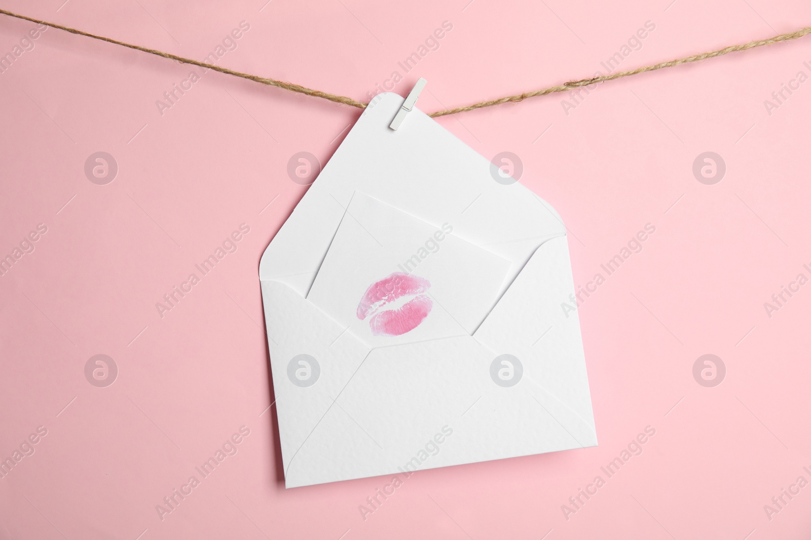 Photo of White envelope and card with lip print hanging on twine against pink background. Love letter