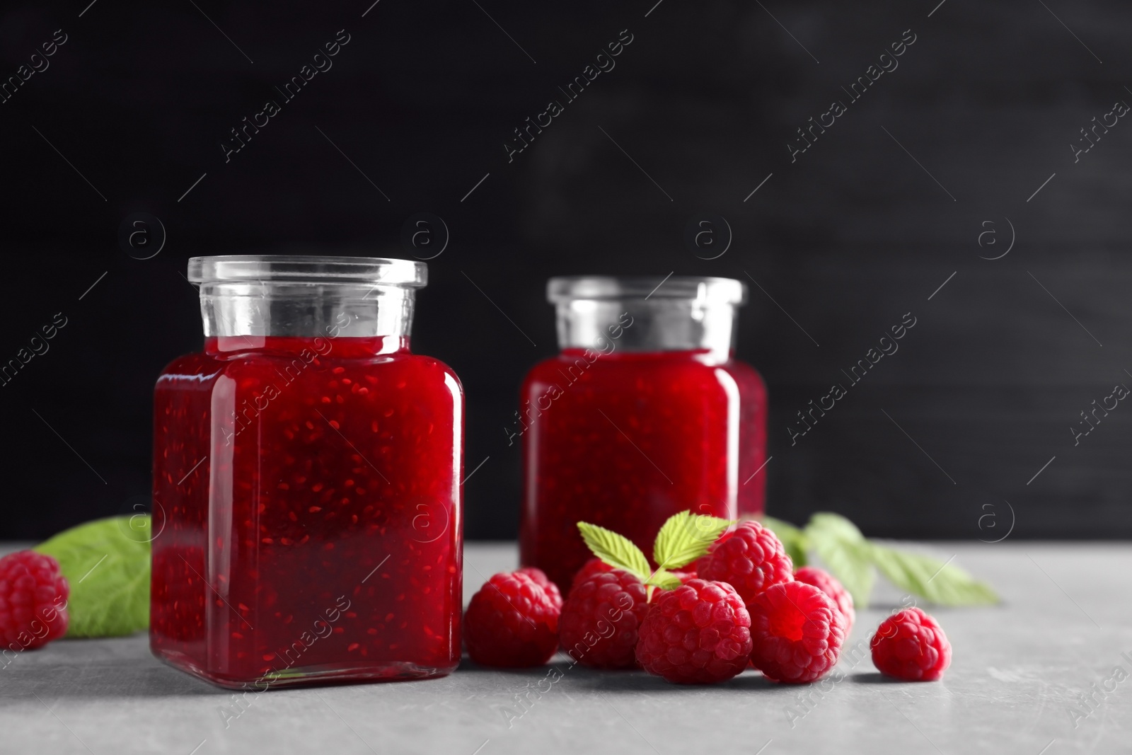 Photo of Glass jars of sweet jam with ripe raspberries and green leaves on table against black background. Space for text