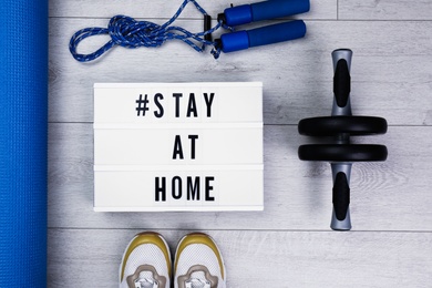 Photo of Sport equipment and lightbox with hashtag STAY AT HOME on floor, flat lay. Message to promote self-isolation during COVID‑19 pandemic