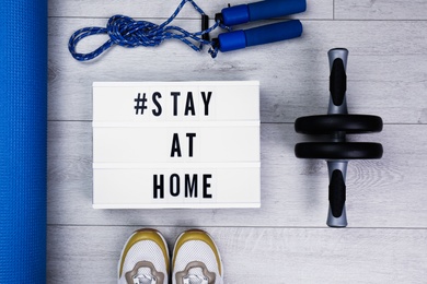 Sport equipment and lightbox with hashtag STAY AT HOME on floor, flat lay. Message to promote self-isolation during COVID‑19 pandemic