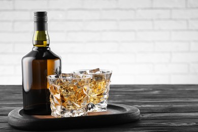 Photo of Whiskey with ice cubes in glasses and bottle on black wooden table, closeup. Space for text