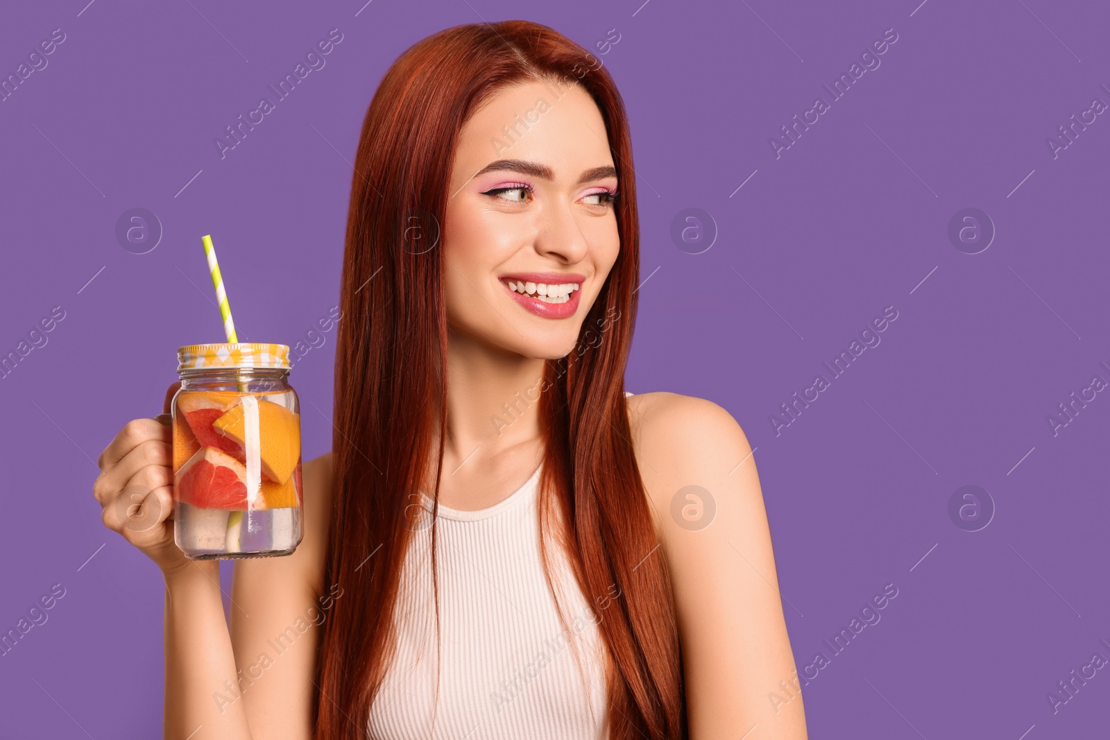 Photo of Happy woman with red dyed hair and refreshing grapefruit drink on purple background