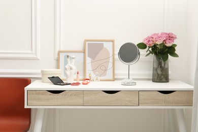 Mirror, cosmetic products, perfumes and vase with pink roses on white dressing table in makeup room