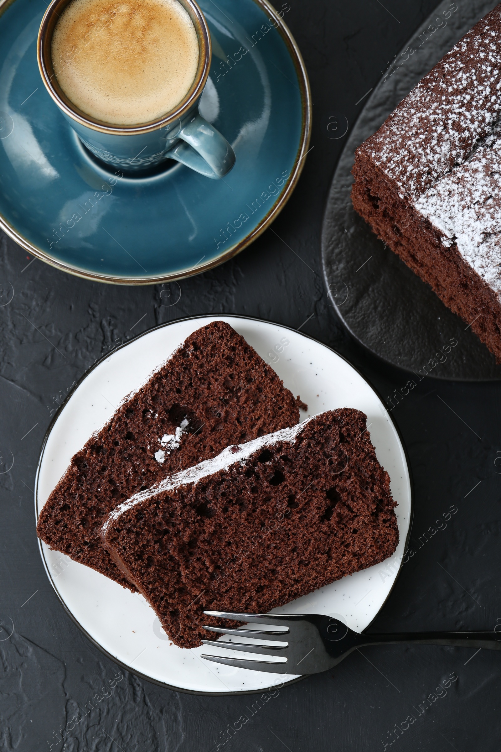 Photo of Tasty chocolate sponge cake with powdered sugar and coffee on black textured table, flat lay