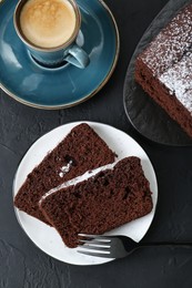 Tasty chocolate sponge cake with powdered sugar and coffee on black textured table, flat lay