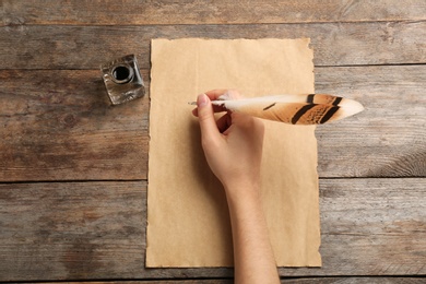 Woman using feather pen to write with ink on parchment at wooden table, top view. Space for text