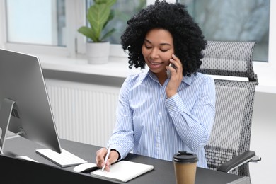 Photo of Young woman talking on phone while working with computer at table in office