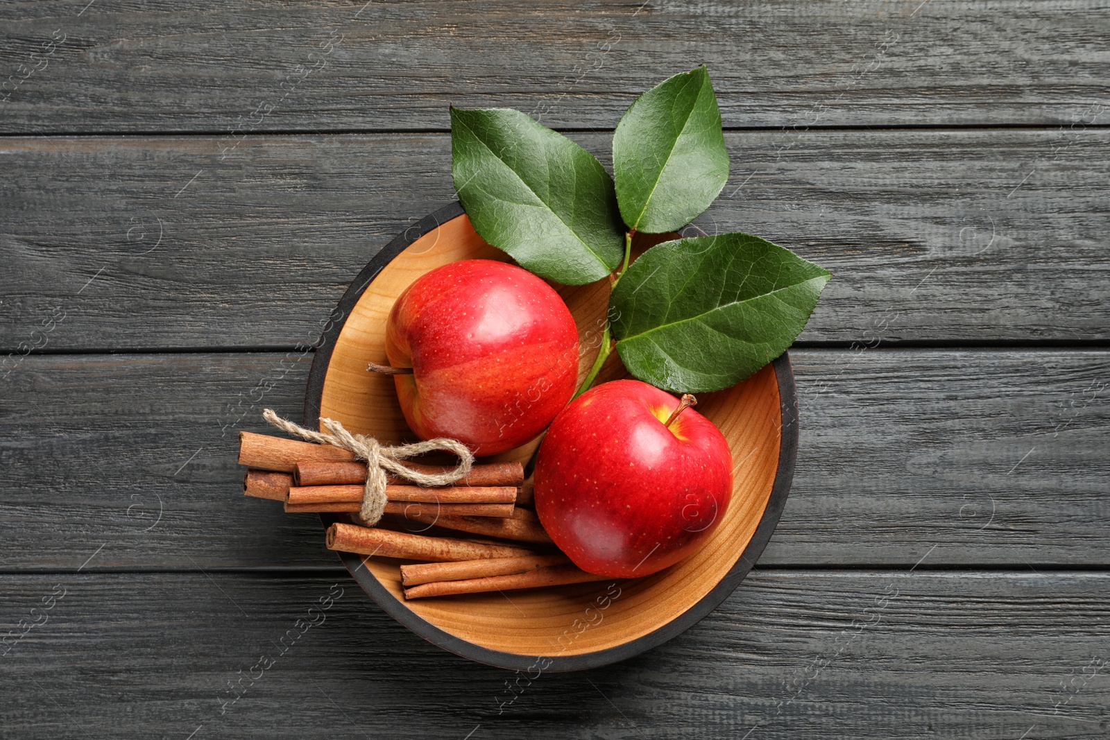 Photo of Fresh apples and cinnamon sticks in bowl on wooden table, top view