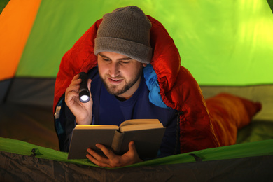 Photo of Young man with flashlight reading book in tent