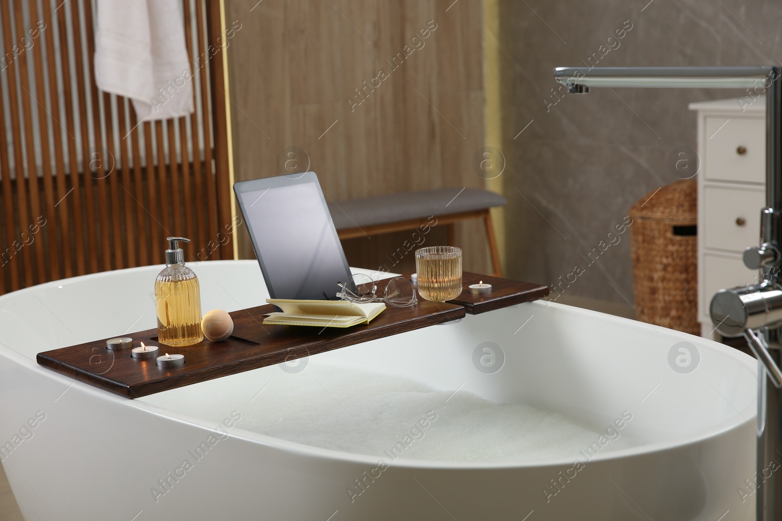 Photo of Wooden bath tray with candles, tablet, book and glass of drink on tub indoors