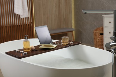 Wooden bath tray with candles, tablet, book and glass of drink on tub indoors