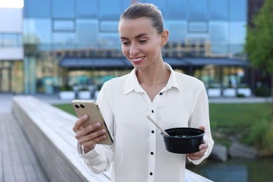 Photo of Smiling businesswoman with lunch box looking at smartphone outdoors
