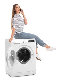 Young woman sitting on washing machine and talking on phone against white background. Laundry equipment