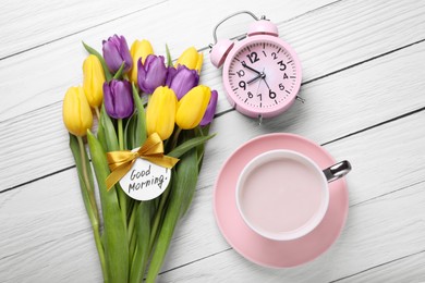 Cup of hot drink, beautiful tulips, alarm clock and card with text Good Morning on white wooden table, flat lay