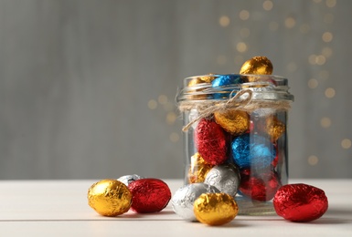 Photo of Glass jar with chocolate eggs wrapped in colorful foil on white wooden table. Space for text