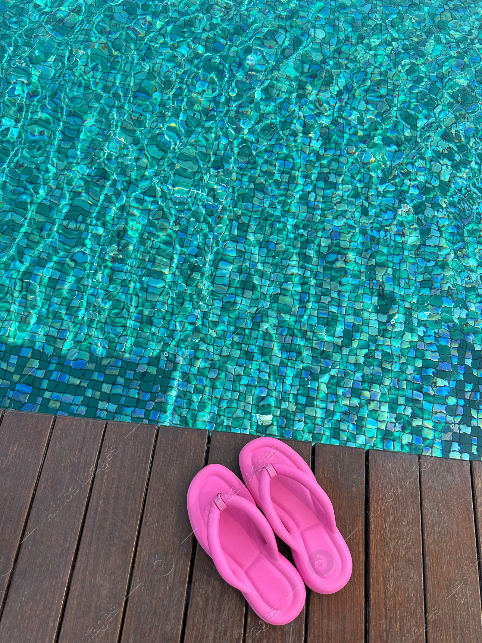 Photo of Clear rippled water in swimming pool and pink flip-flops on wooden deck outdoors, above view. Space for text