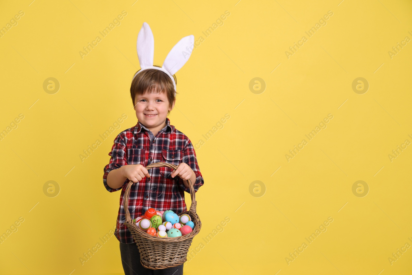 Photo of Cute little boy wearing bunny ears with basket full of dyed Easter eggs on yellow background, space for text