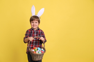 Photo of Cute little boy wearing bunny ears with basket full of dyed Easter eggs on yellow background, space for text