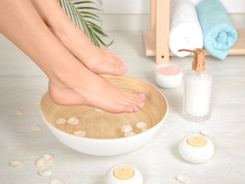 Photo of Woman putting her feet into bowl with water and rose petals indoors, closeup. Spa treatment