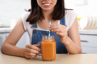 Woman with mason jar of tasty carrot juice at table indoors, closeup