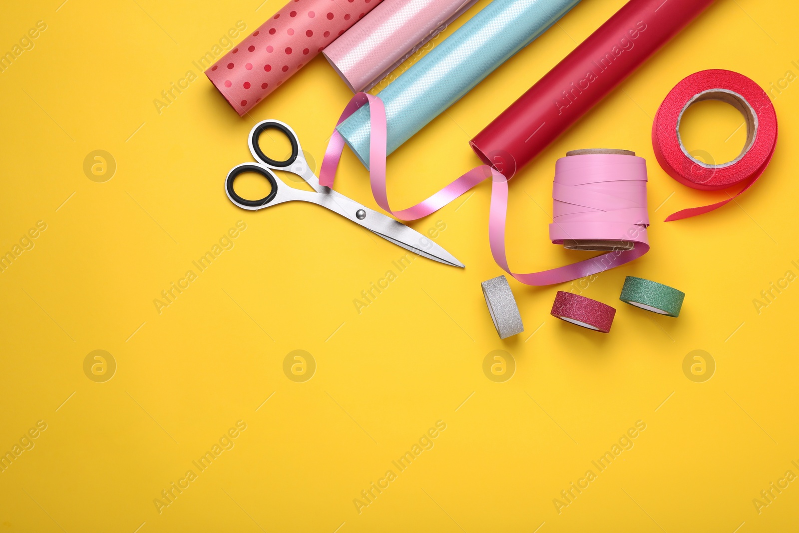 Photo of Rolls of colorful wrapping papers, scissors and ribbons on yellow background, flat lay. Space for text