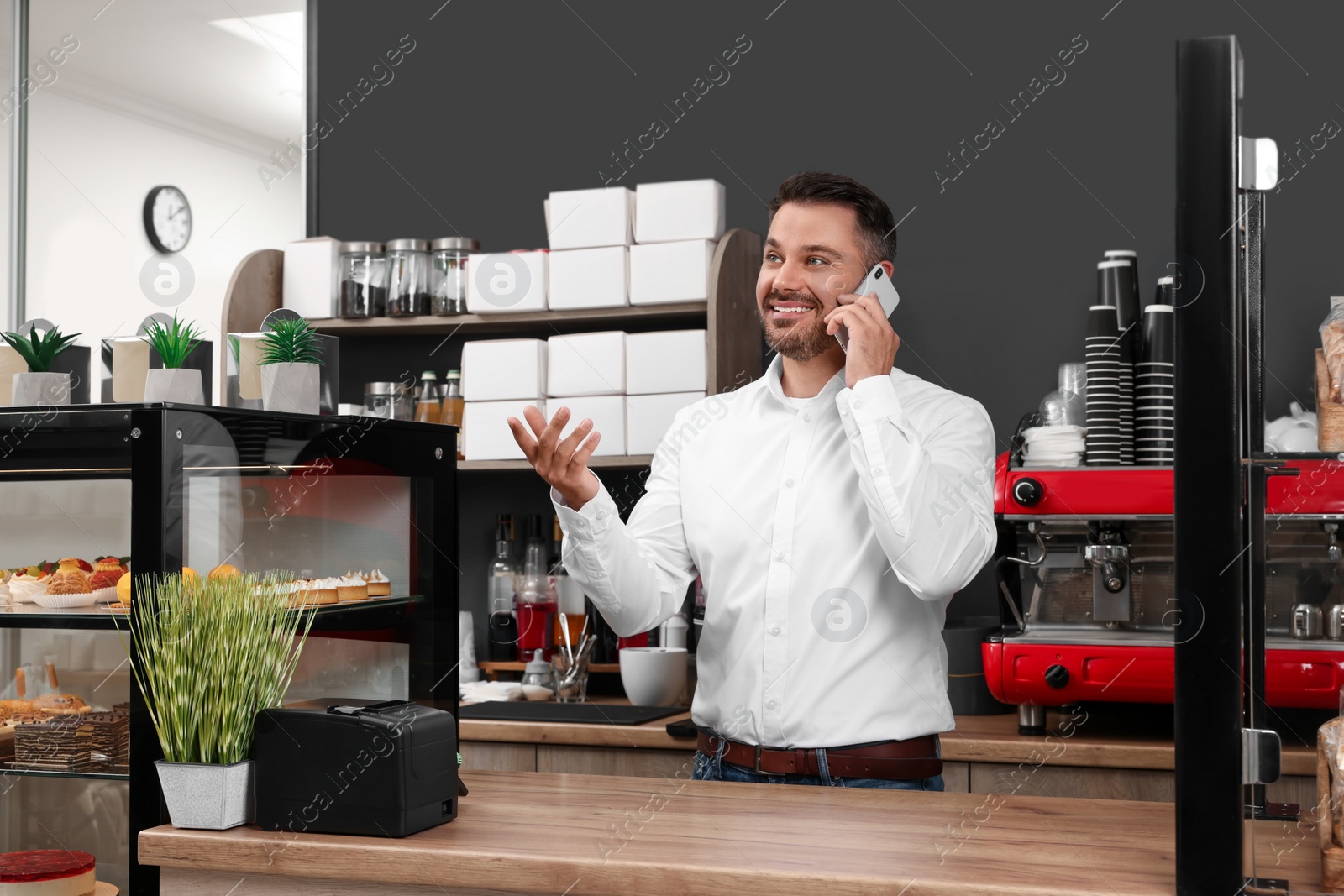 Photo of Business owner in his cafe. Man talking on phone while using tablet near showcase with pastries