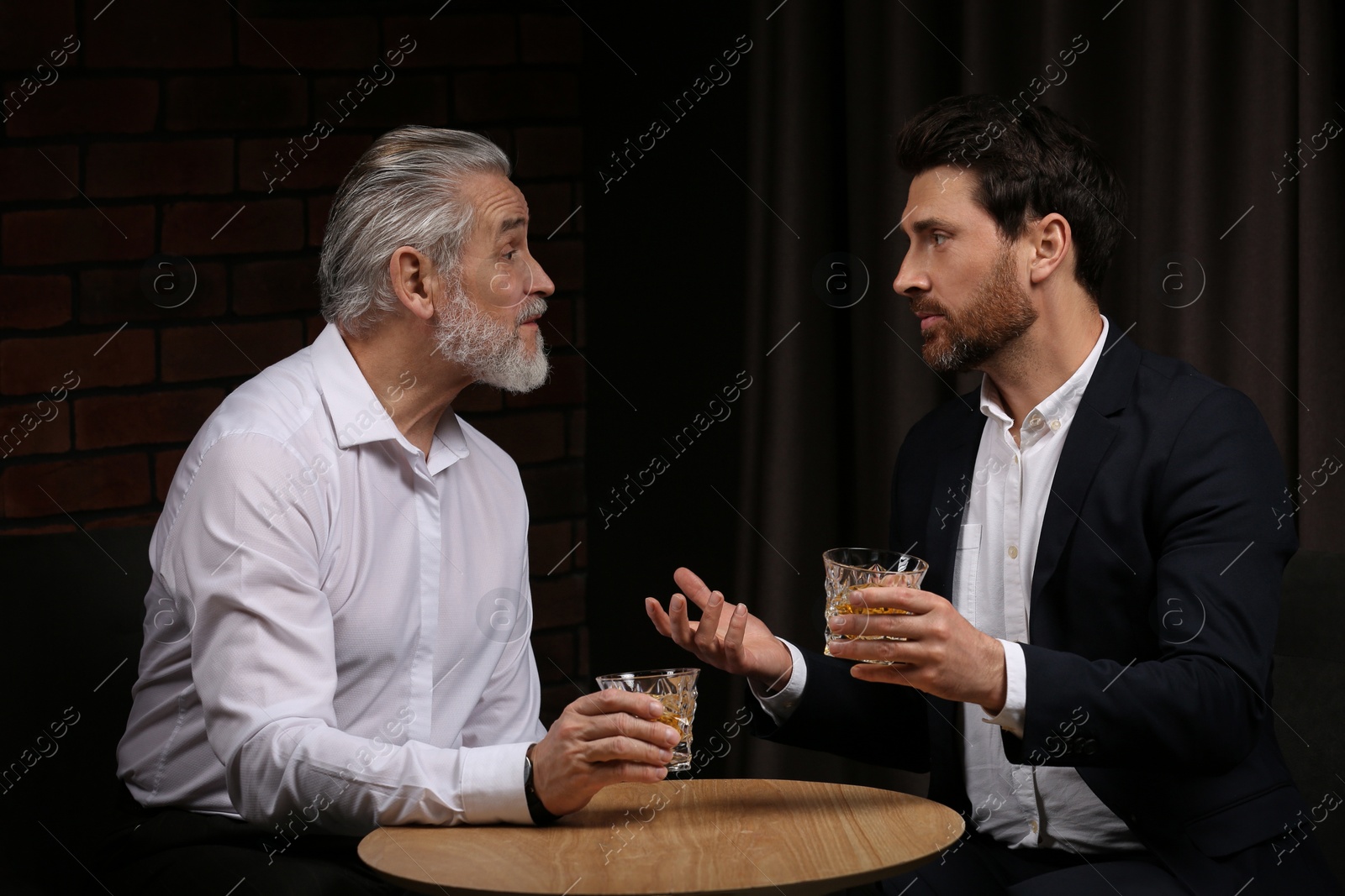 Photo of Men with glasses of whiskey talking at wooden table indoors