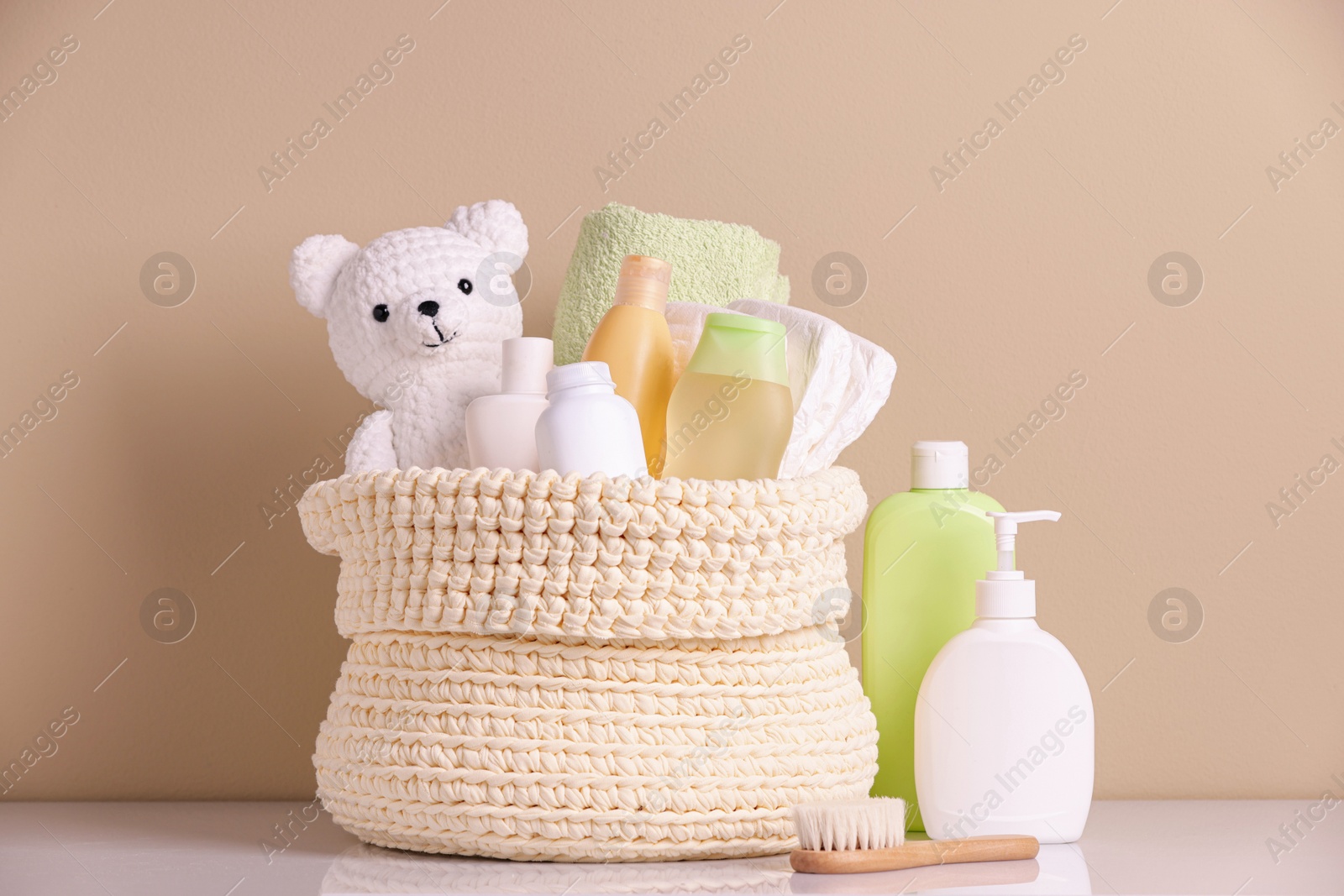 Photo of Knitted basket with baby cosmetic products and toy bear on white table against beige background