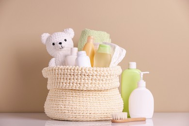 Photo of Knitted basket with baby cosmetic products and toy bear on white table against beige background