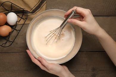 Photo of Woman whipping ingredients with balloon whisk at wooden table, top view. Cooking delicious eggnog