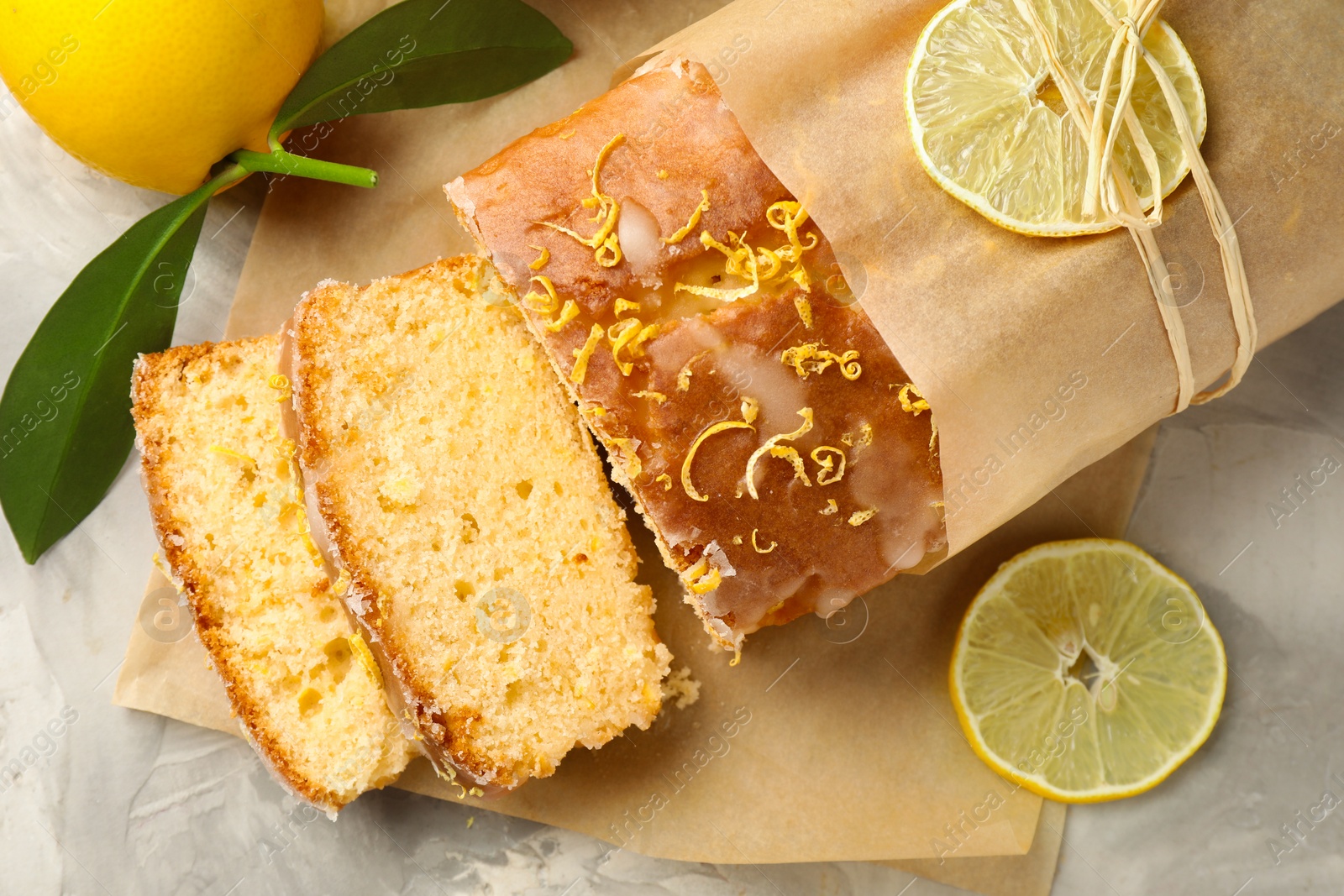 Photo of Cut tasty lemon cake with glaze and citrus fruits on light grey textured table, flat lay