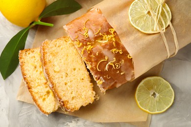 Photo of Cut tasty lemon cake with glaze and citrus fruits on light grey textured table, flat lay