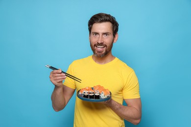 Happy man holding plate with tasty sushi rolls and chopsticks on light blue background