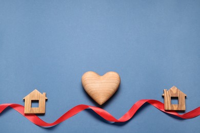 Photo of Decorative heart and red ribbon between two wooden house models on blue background symbolizing connection in long-distance relationship, flat lay. Space for text