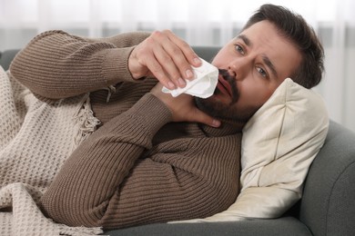 Sick man with tissue blowing nose on sofa indoors. Cold symptoms