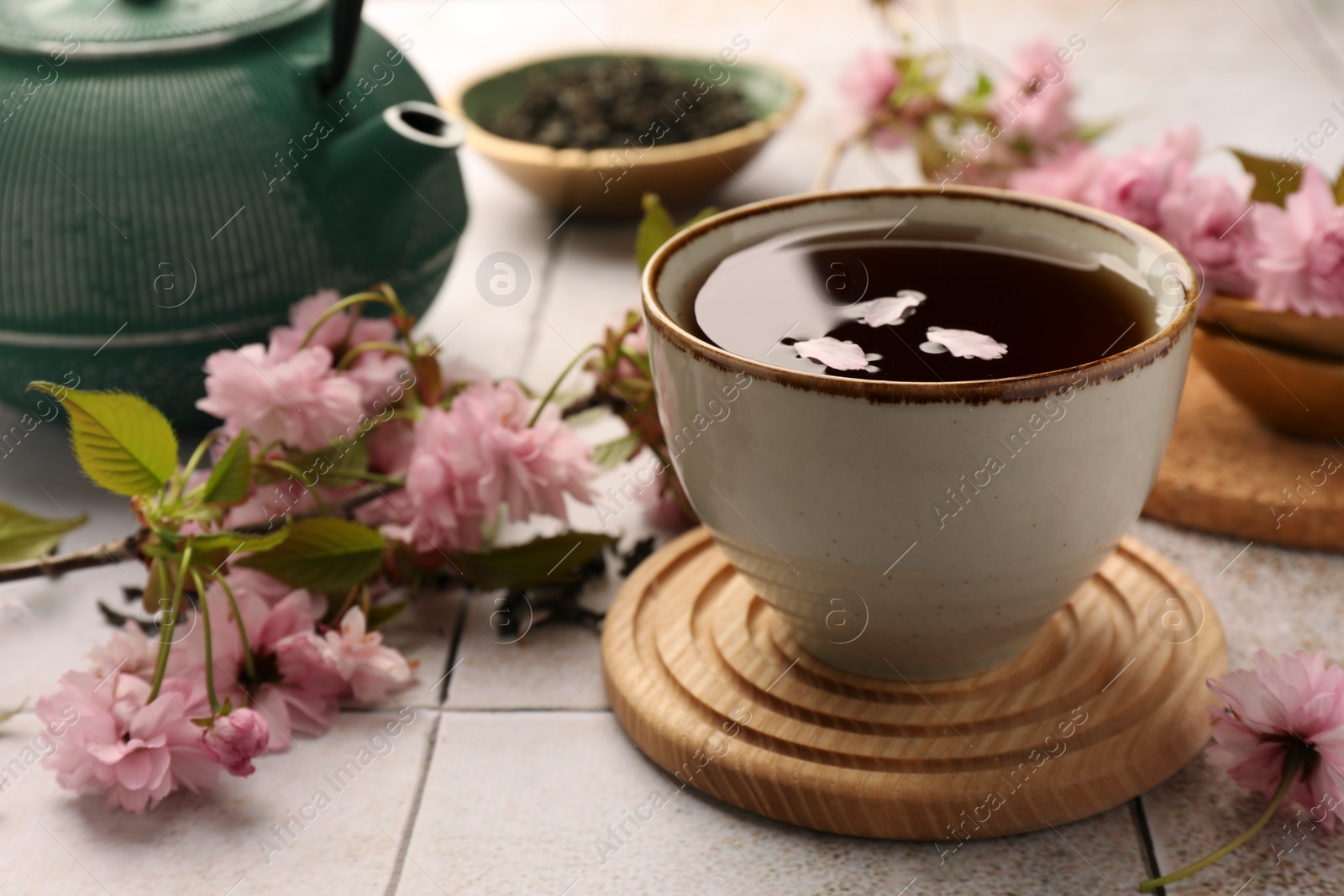 Photo of Traditional ceremony. Sakura petals in cup of brewed tea, teapot and flowers on tiled table, closeup