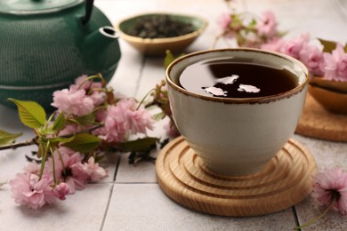 Traditional ceremony. Sakura petals in cup of brewed tea, teapot and flowers on tiled table, closeup