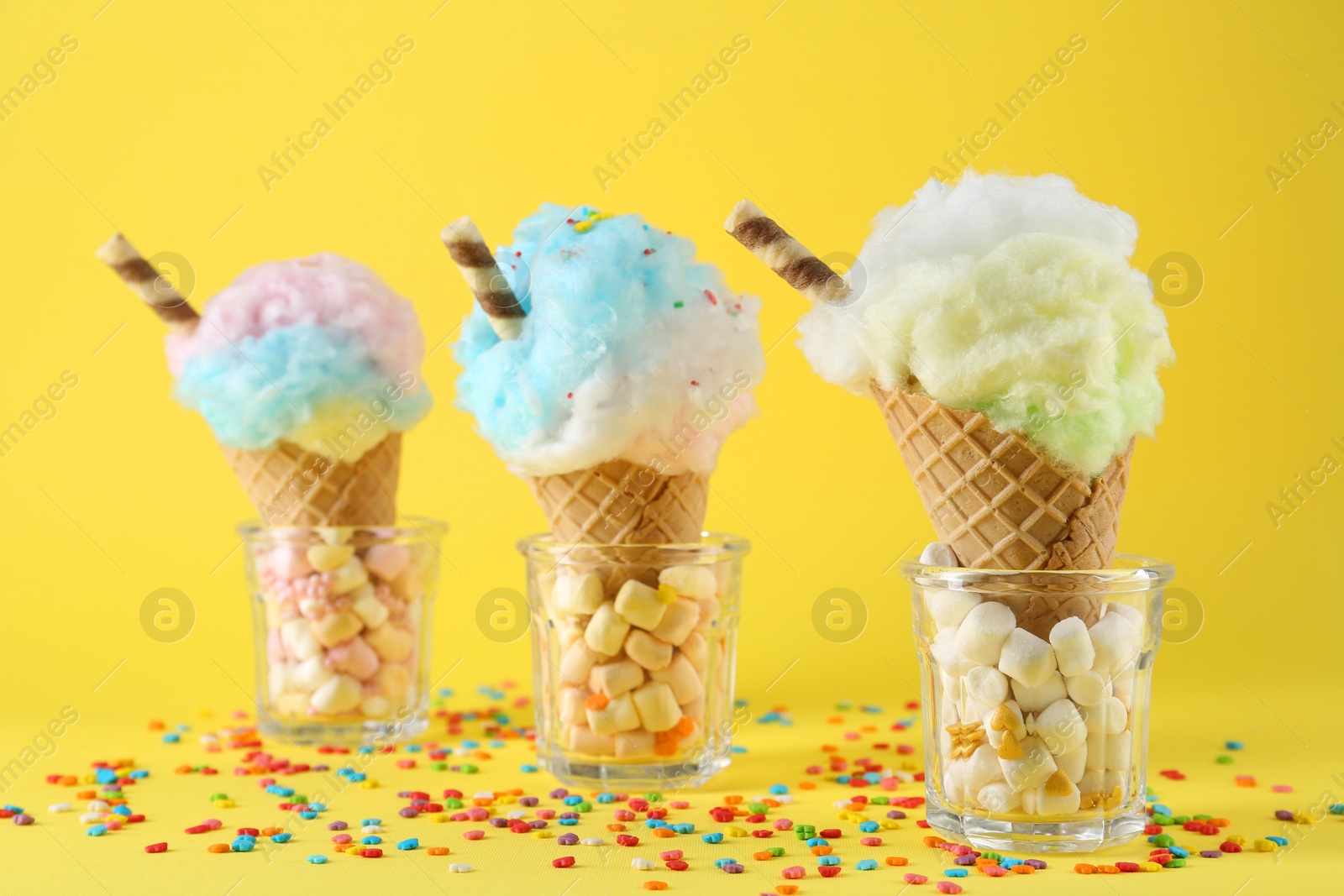 Photo of Sweet cotton candies in waffle cones and marshmallows on yellow background, closeup