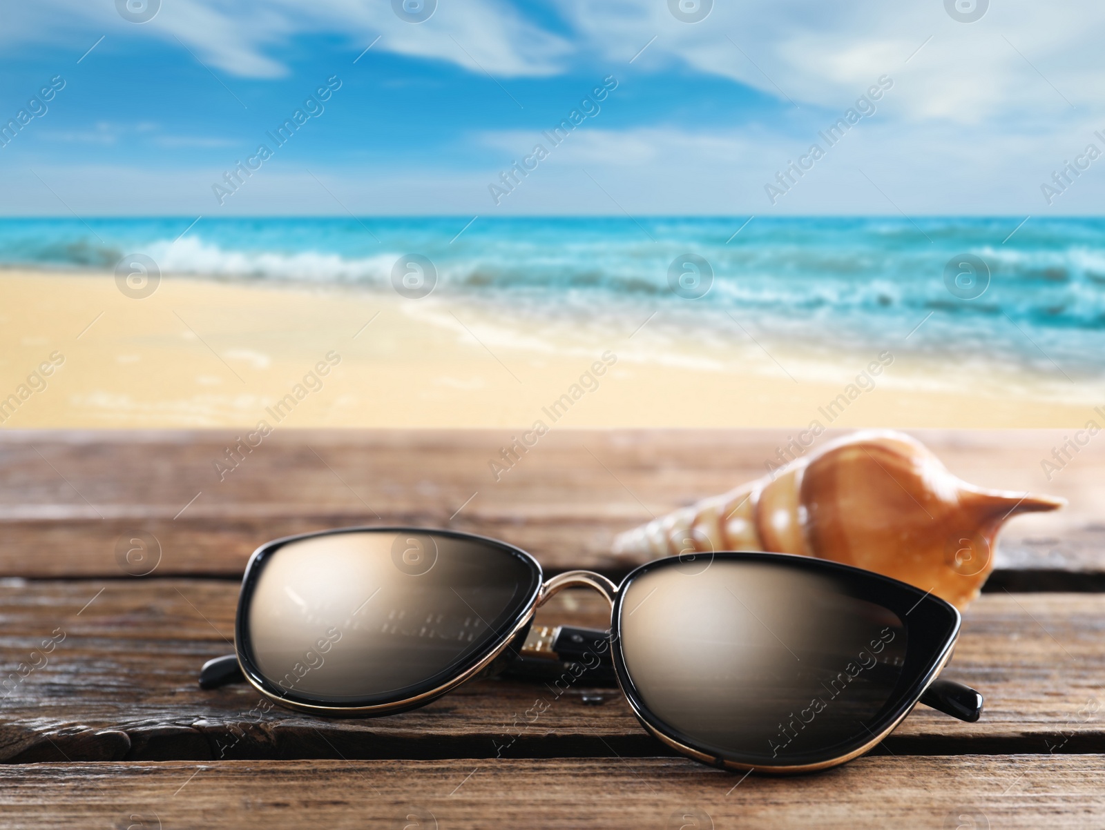 Image of Shell and stylish sunglasses on wooden table near sea with sandy beach