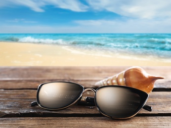 Image of Shell and stylish sunglasses on wooden table near sea with sandy beach