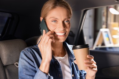 Photo of Coffee to go. Happy woman with paper cup of drink talking on smartphone in car