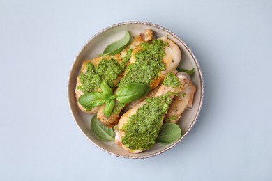 Delicious fried chicken drumsticks with pesto sauce and basil in bowl on light gray table, top view