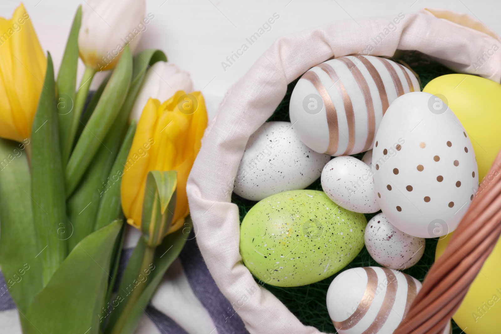 Photo of Easter eggs in basket and tulips on white table, top view