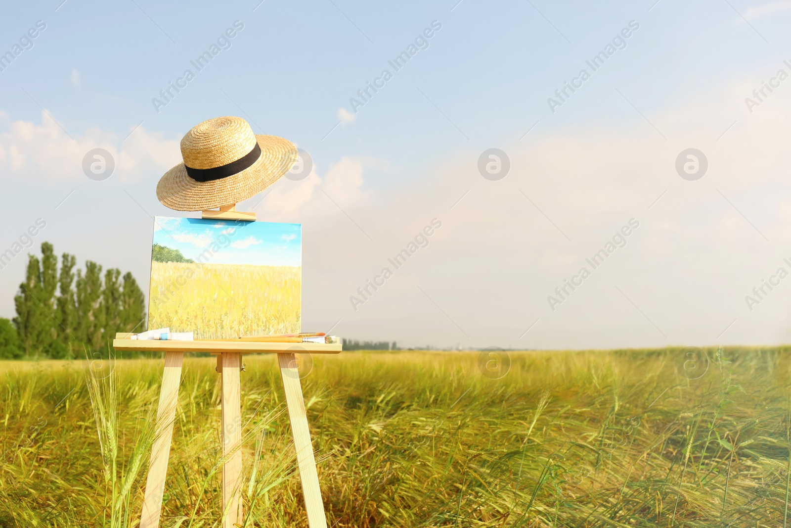 Photo of Wooden easel with beautiful picture, painting equipment and hat in field. Space for text