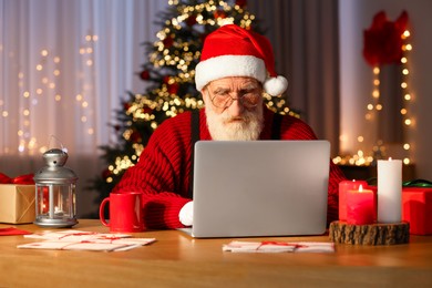 Santa Claus using laptop at his workplace in room with Christmas tree